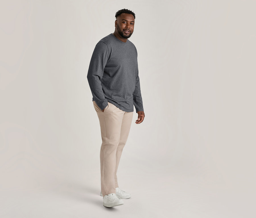 Homme souriant portant un t-shirt gris foncé à manches longues, un pantalon chino beige et des baskets blanches, une main dans la poche.