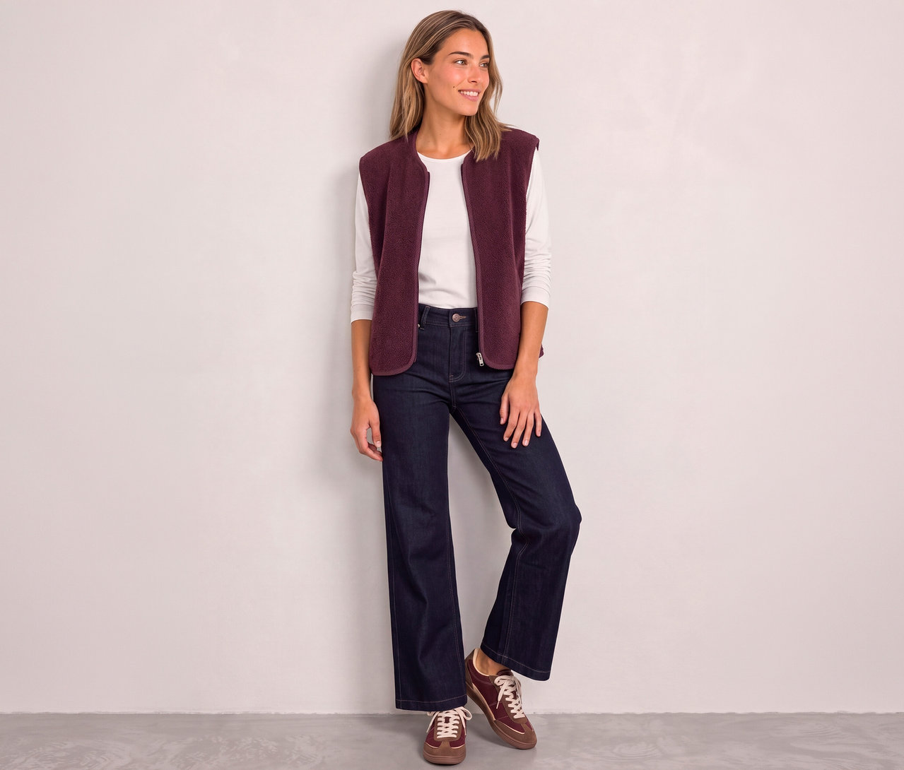 Une femme souriante porte un gilet Street One rouge sur un t-shirt blanc à manches longues, un jean large et des baskets marron.