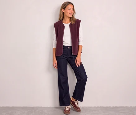 Une femme souriante portant un gilet Street One rouge sur un haut blanc à manches longues, un jean foncé et des baskets bordeaux, est appuyée contre un mur blanc.