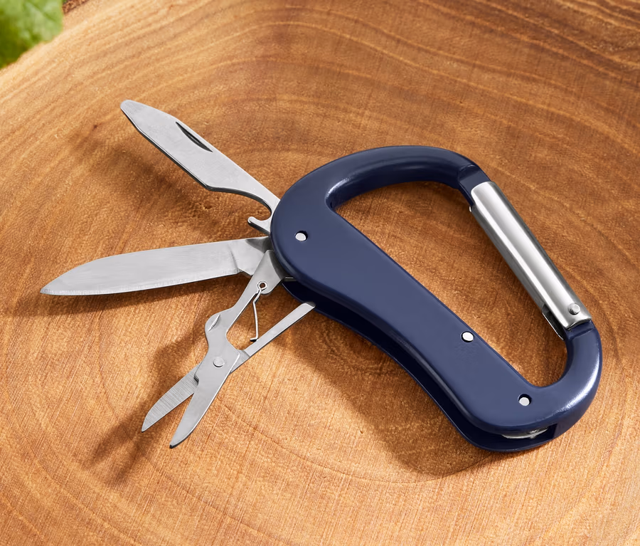 Un mousqueton avec outils polyvalents 5 en 1 avec couteau, scie et ciseaux posé sur une souche d'arbre.