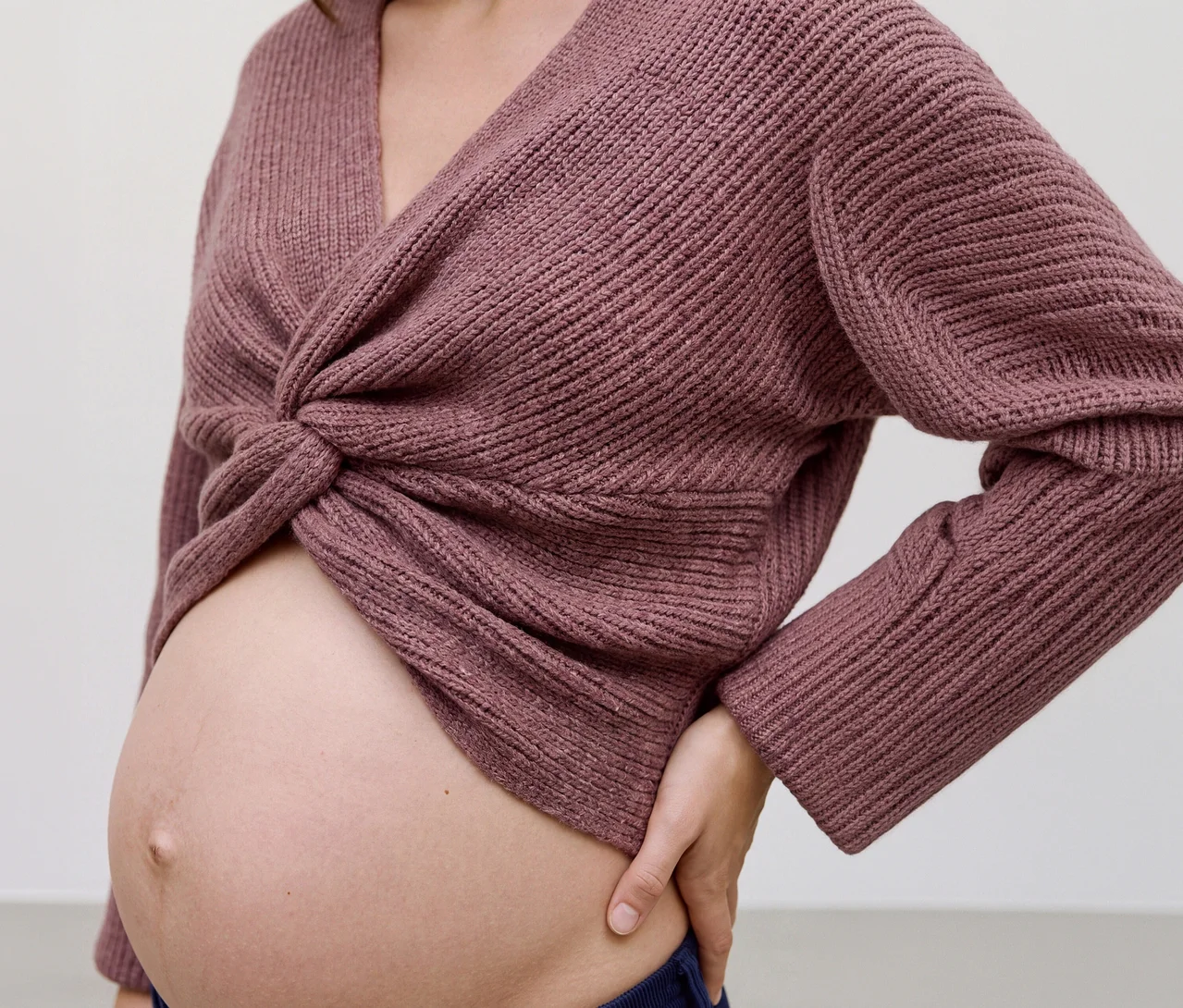 Une femme enceinte porte un haut court en maille MAMALICIOUS « SVALA » de couleur mauve avec un devant torsadé, exposant son ventre. Sa main gauche repose sur sa hanche.
