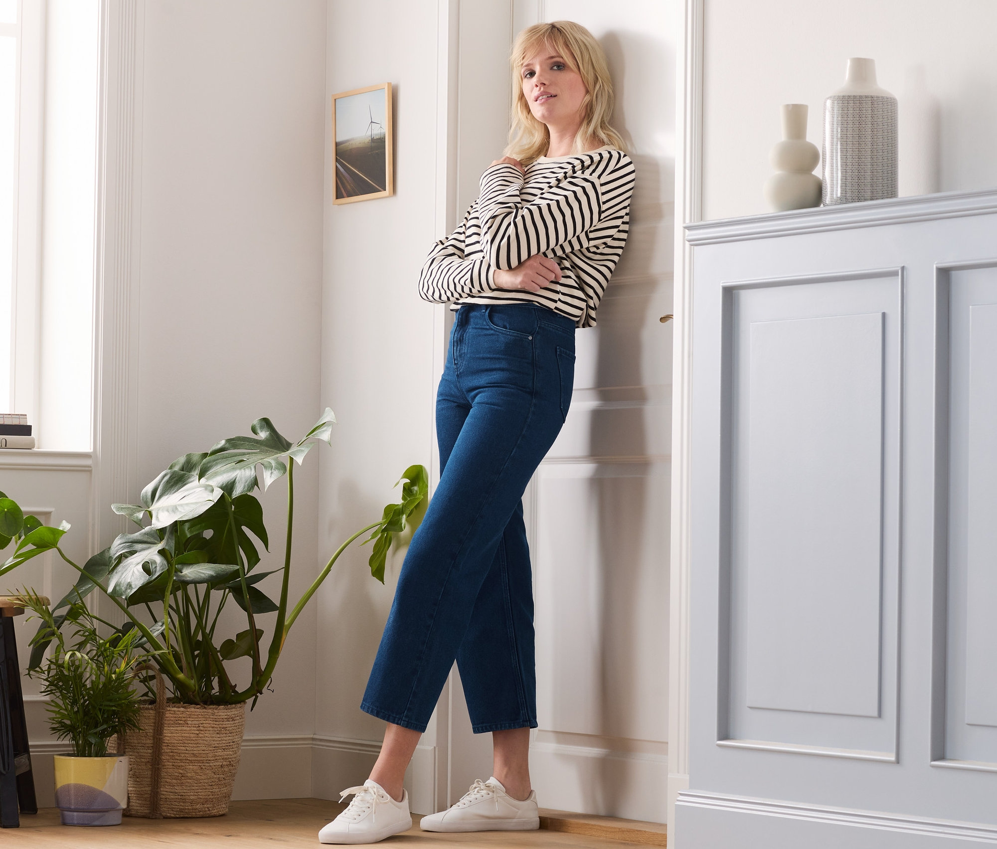 Une femme aux bras croisés est appuyée contre une porte et porte un jean large, un haut rayé et des baskets.