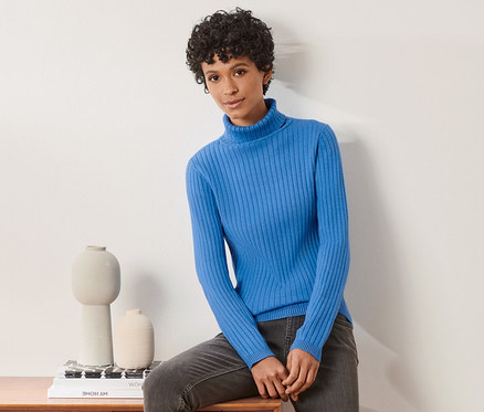 Une femme est assise sur une commode en bois et porte un pull à col roulé côtelé bleu.