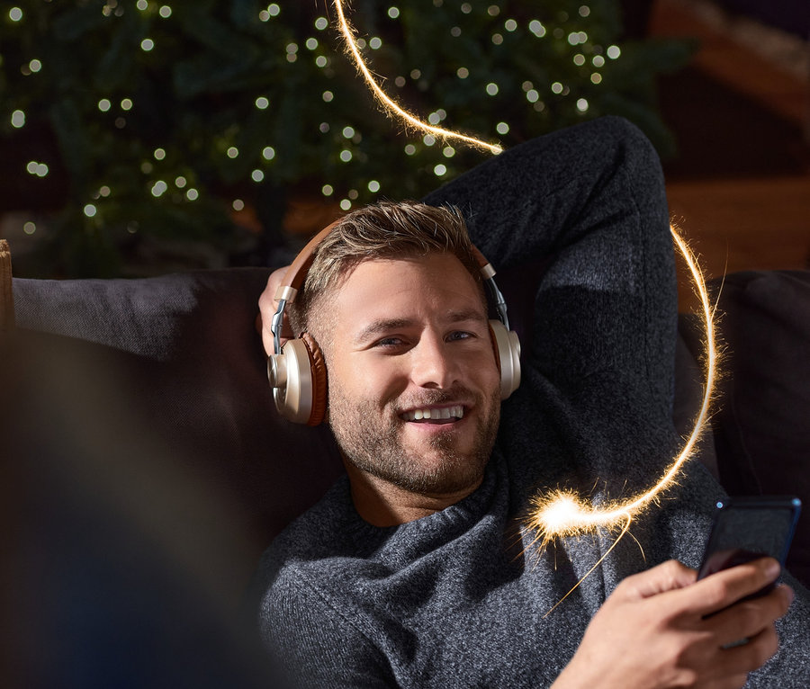 Un homme souriant allongé sur un canapé avec un casque Bluetooth® sur les oreilles tient un téléphone à la main. Un sapin de Noël est visible en arrière-plan.