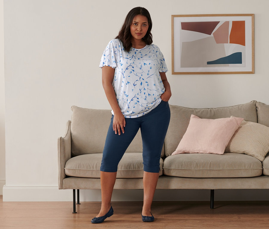 Une femme se tient dans une pièce et porte un chemisier blanc à manches chauve-souris, un corsaire bleu foncé en bengaline et des ballerines en cuir.