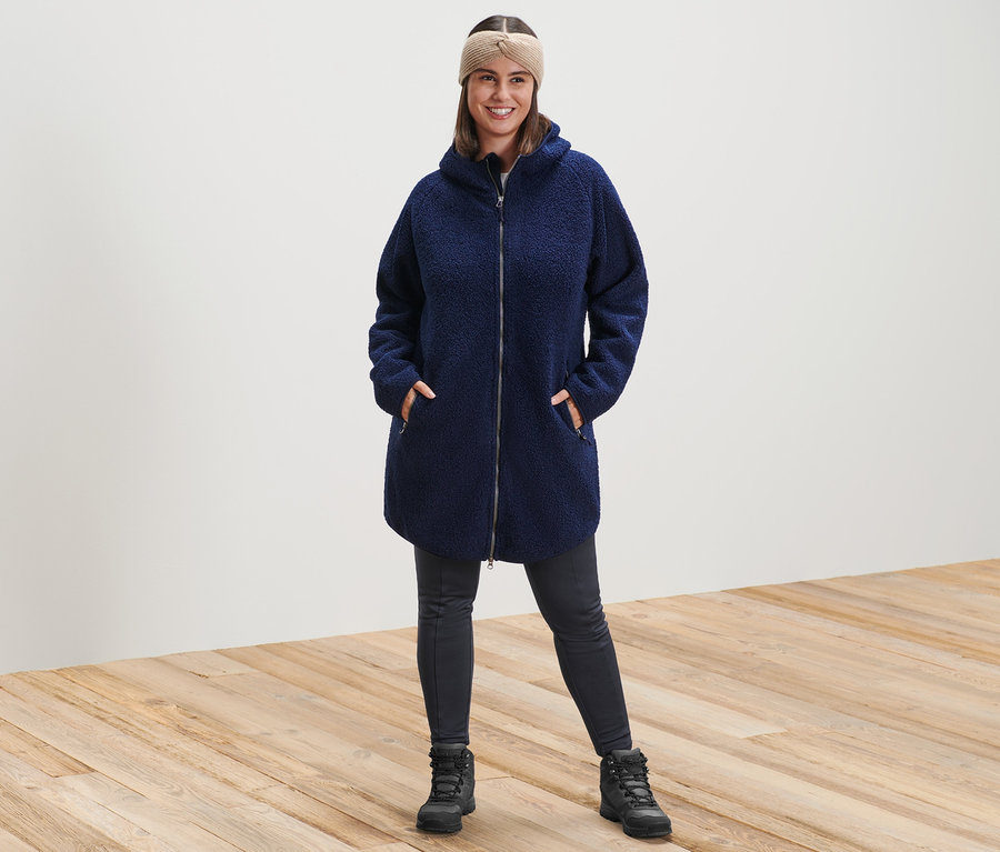 Une femme debout porte un manteau teddy à capuche, un bandeau en maille couleur sable et un legging thermique.