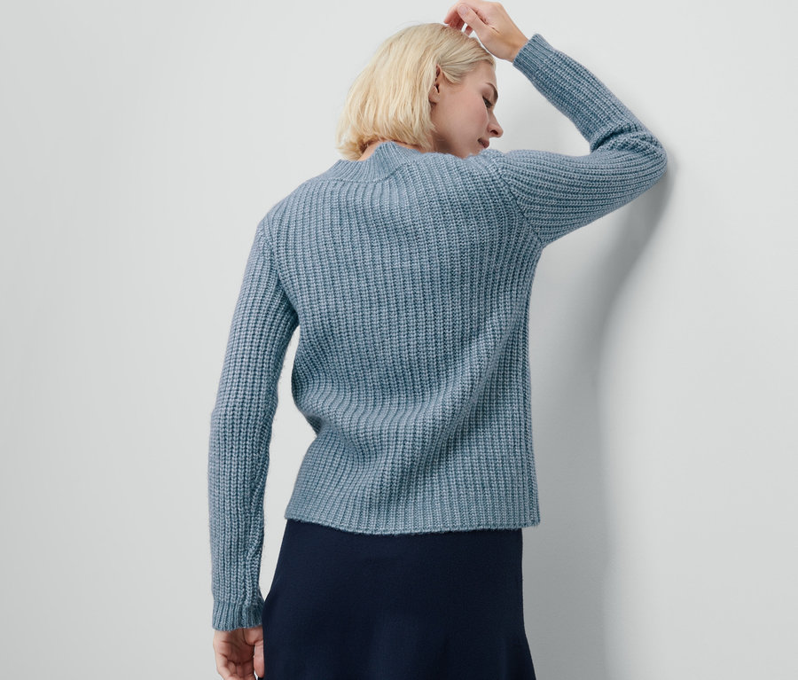 Une femme blonde a une main sur la tête et porte un cardigan en grosse maille bleu clair.