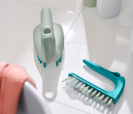 Deux brosses à sols et carrelage 2 en 1 sont posées sur des carreaux blancs à côté d'un lavabo.