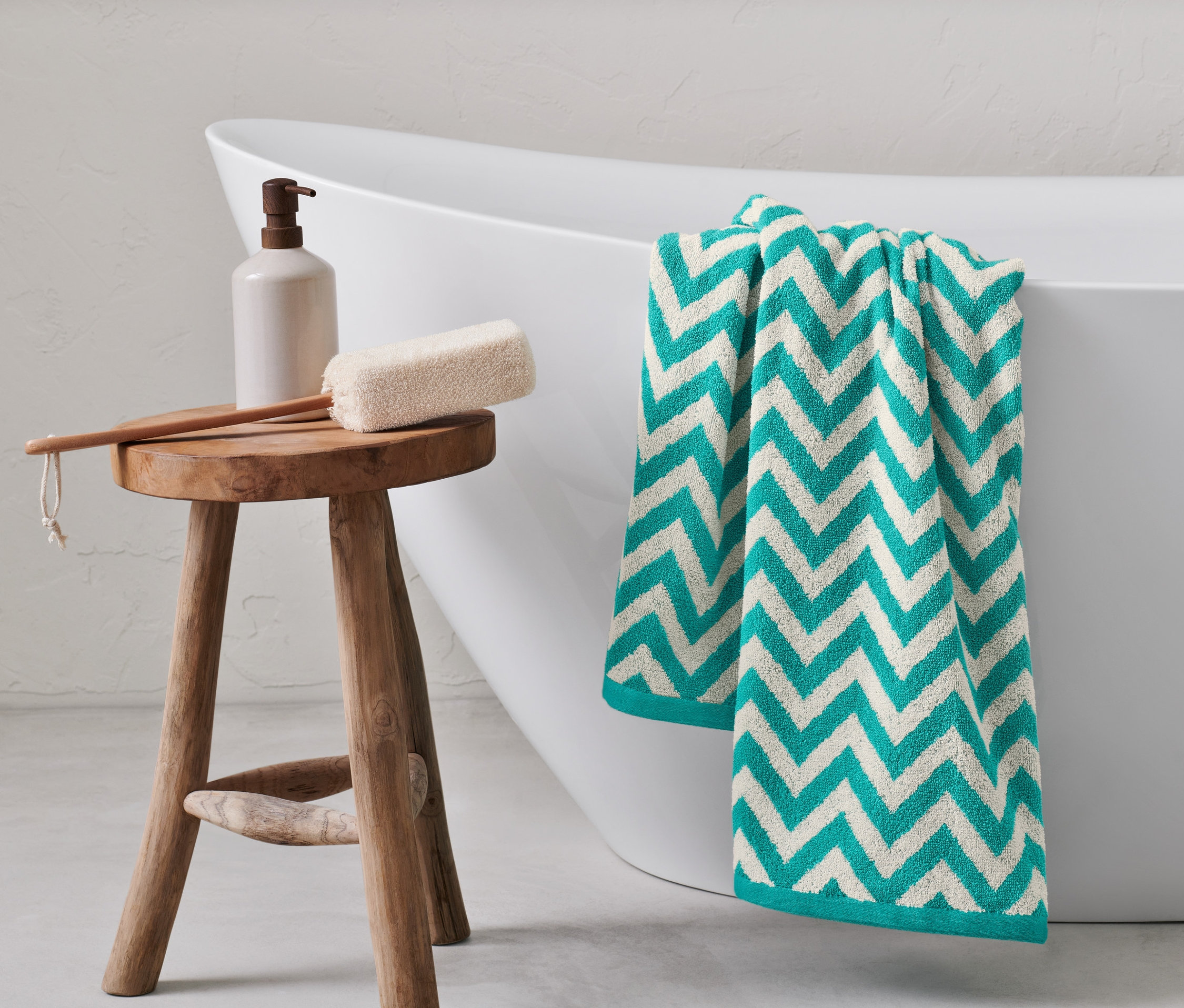 Un drap de bain en jacquard turquoise est suspendu au bord d'une baignoire blanche. Un tabouret en bois supporte un distributeur de savon, une éponge de bain et une brosse.
