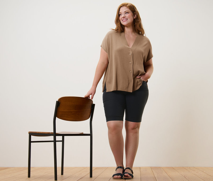 Une femme se tient à côté d'une chaise et porte un cardigan beige à manches courtes et un bermuda en bengaline.