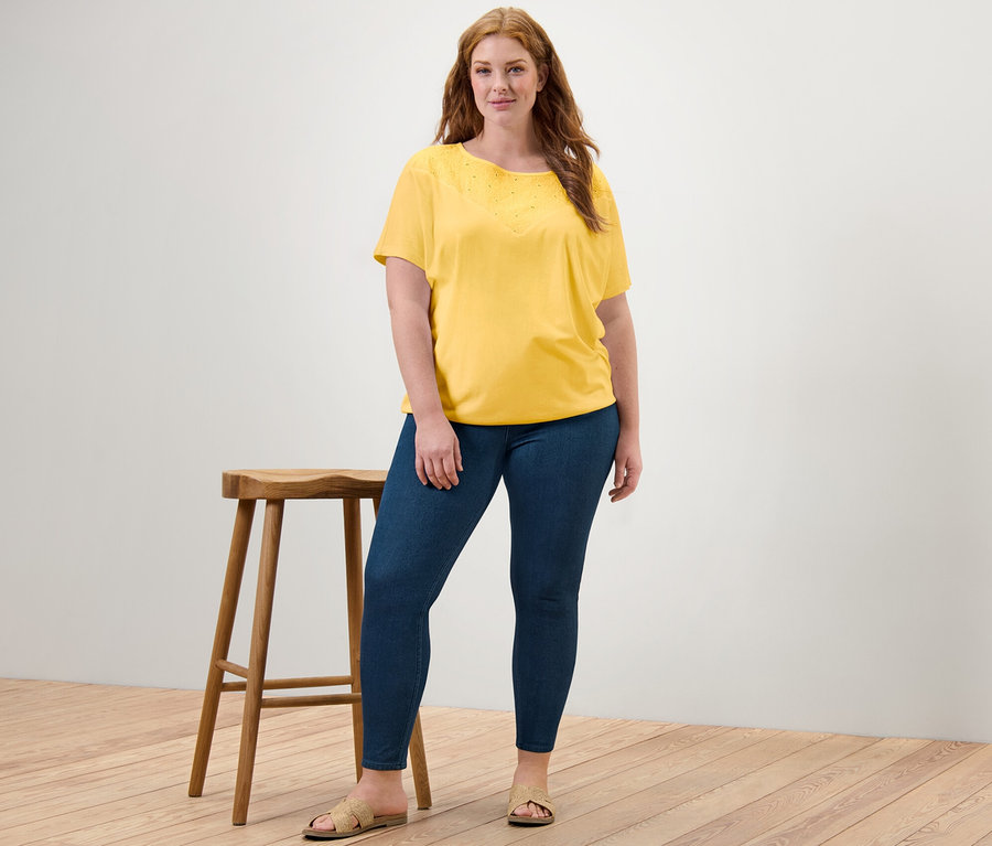 Femme posant à côté d'un tabouret portant un chemisier jaune avec broderie et un jegging.