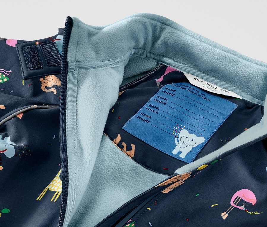 Gros plan sur une combinaison en softshell pour enfant avec des motifs d'animaux, fermeture éclair et étiquette de nom visible.