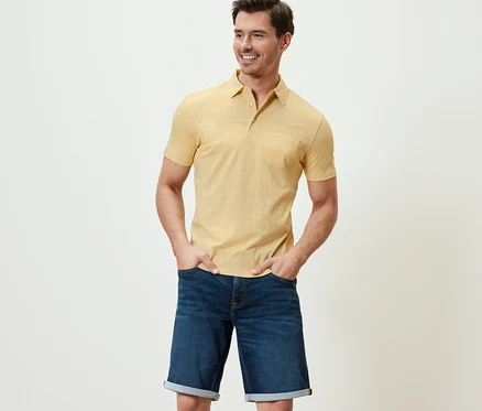 Un homme pose en polo en jersey jaune chiné et short en jean.