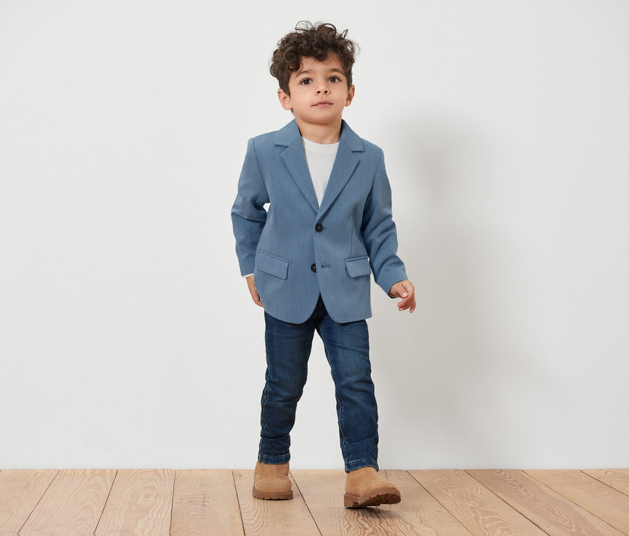 Garçon portant un veston pour enfant et se tenant sur un plancher en bois.