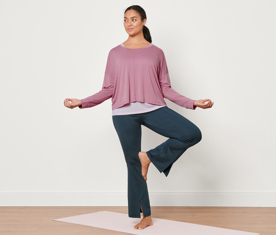 Femme en posture de l'arbre sur un tapis de yoga portant un pantalon de sport modelant bleu foncé et un t-shirt de sport 2 en 1 rose.