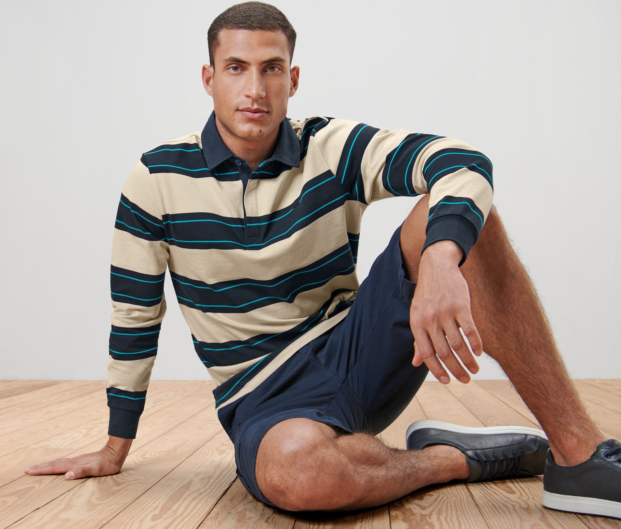 Un homme est assis sur un plancher en bois et porte un polo à manches longues, un short et des baskets.