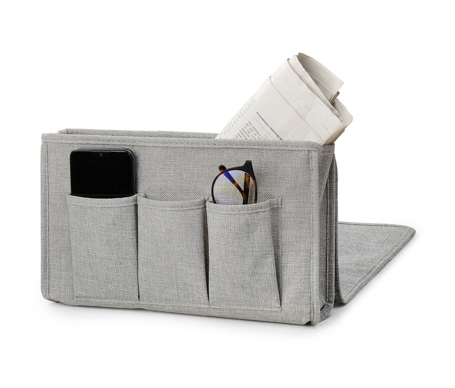 Un rangement de lit ou canapé gris avec des poches contenant un téléphone, des lunettes et un journal.