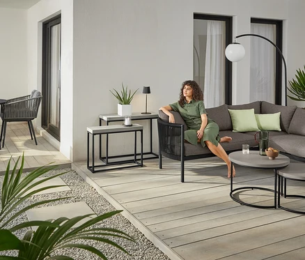 Une femme est assise sur le coin lounge « Jara » avec tressage textile à côté de 2 tables basses avec plateau de table en béton. À l'arrière plan, une table de jardin ronde « Jara » en Duraboard.
