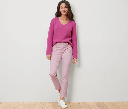 Une femme debout porte un jean coloré rose – Coupe « Emma » et un pull violet.