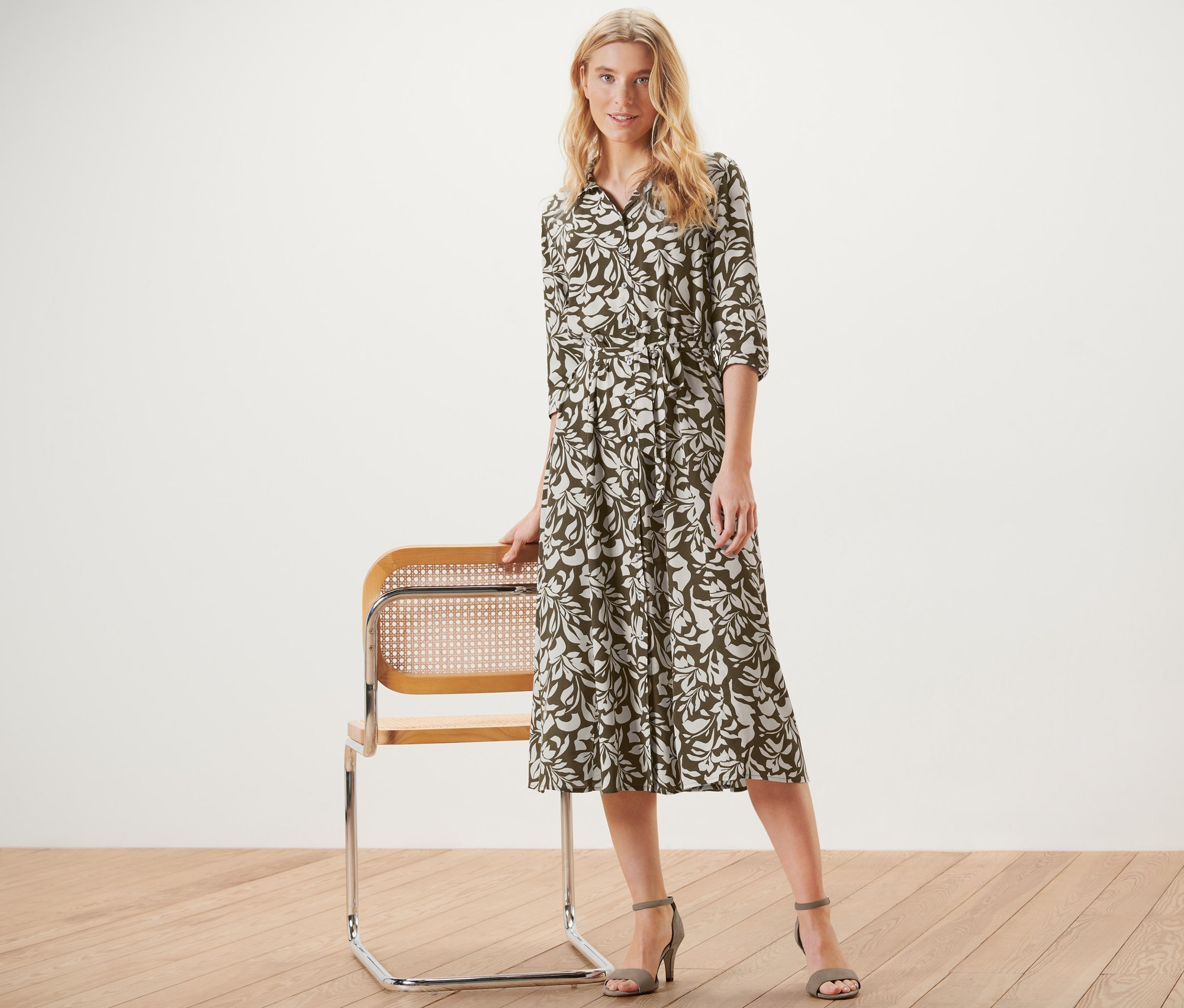 Une femme blonde pose à côté d'une chaise et porte une robe chemise imprimée.