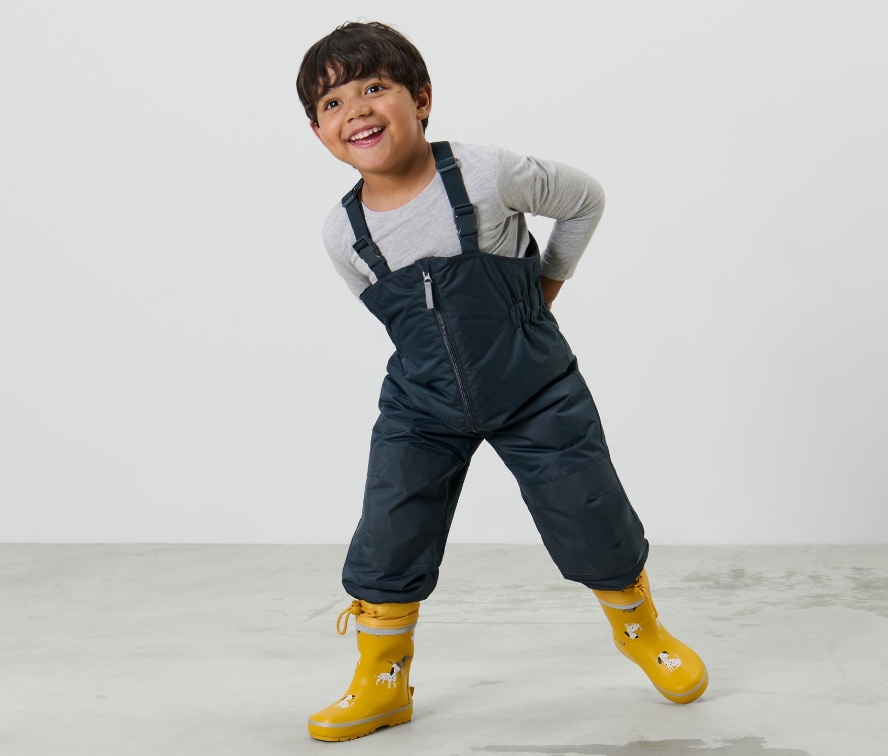 Un garçon pose en pantalon de pluie évolutif pour enfant et bottes en caoutchouc jaunes.