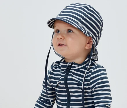 Un bébé porte un bonnet d’été à rayures avec un indice de protection UV 80 et une combinaison rayée.