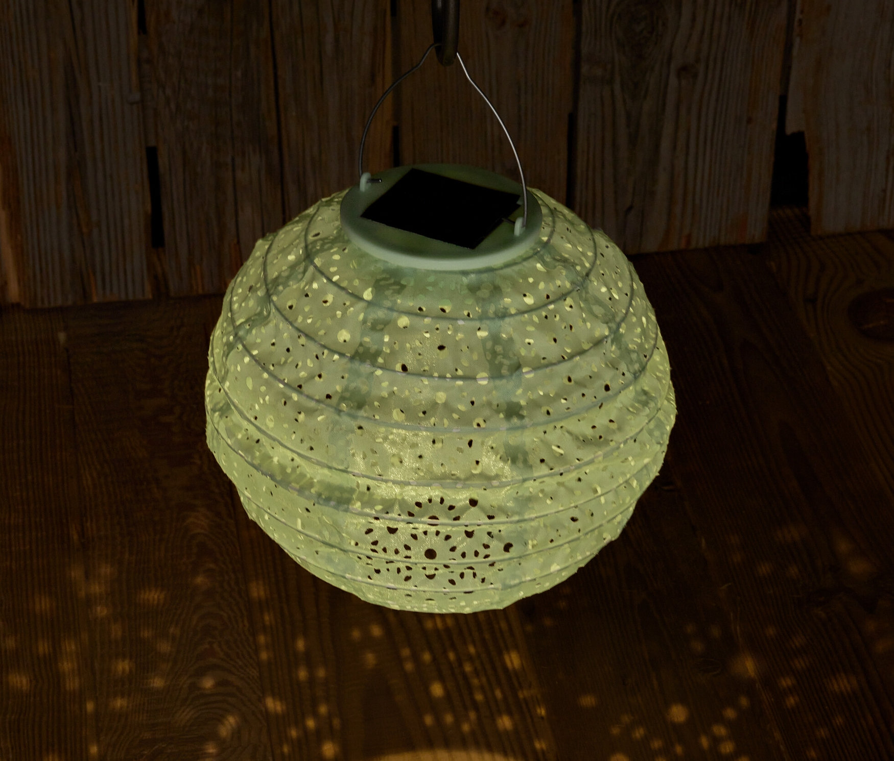 Un lampion solaire suspendu avec des trous décoratifs projette de la lumière sur un plancher en bois.