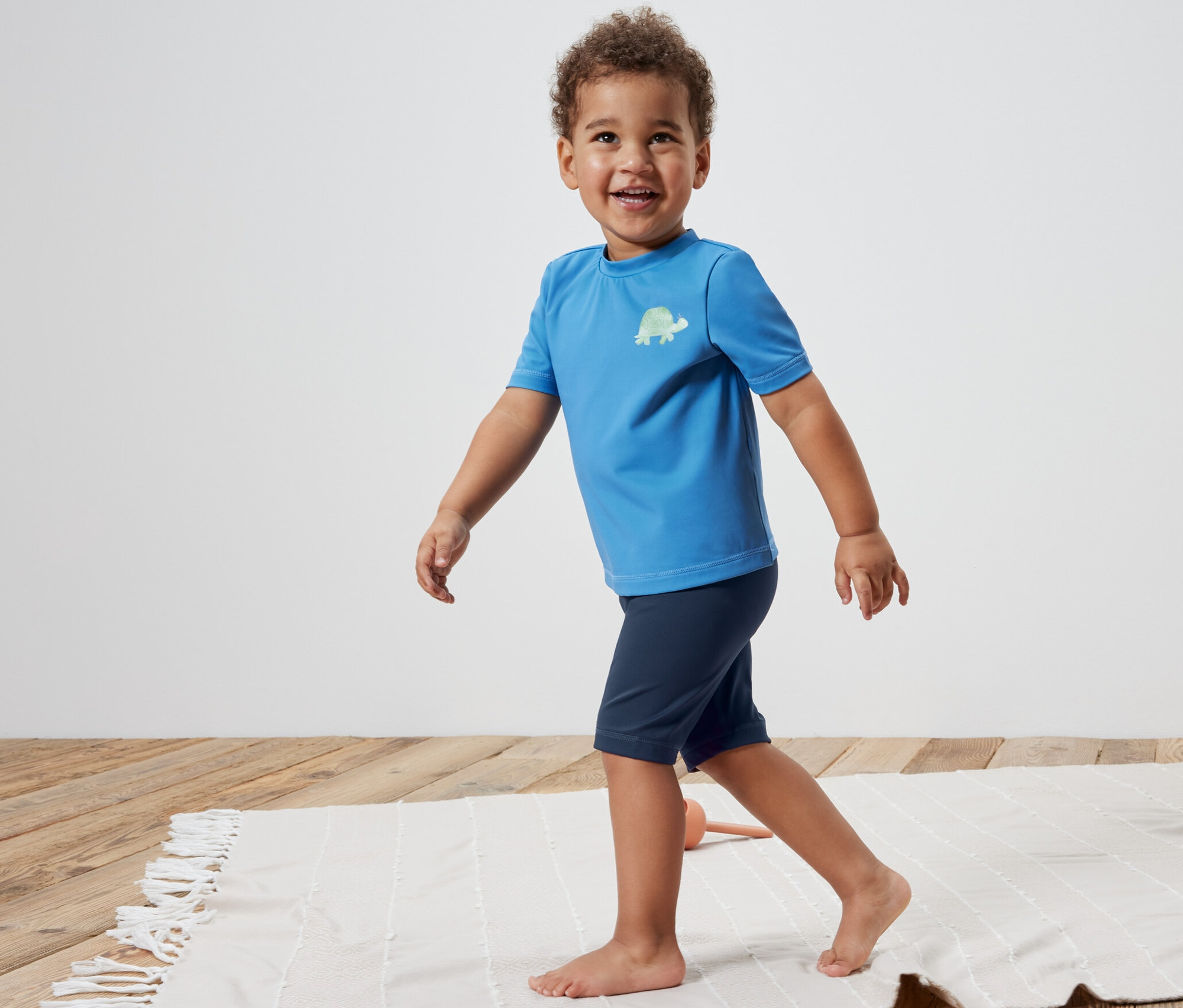 Un garçon marche sur la plage en portant un maillot de bain de natation pour enfant avec indice de protection UV 80.