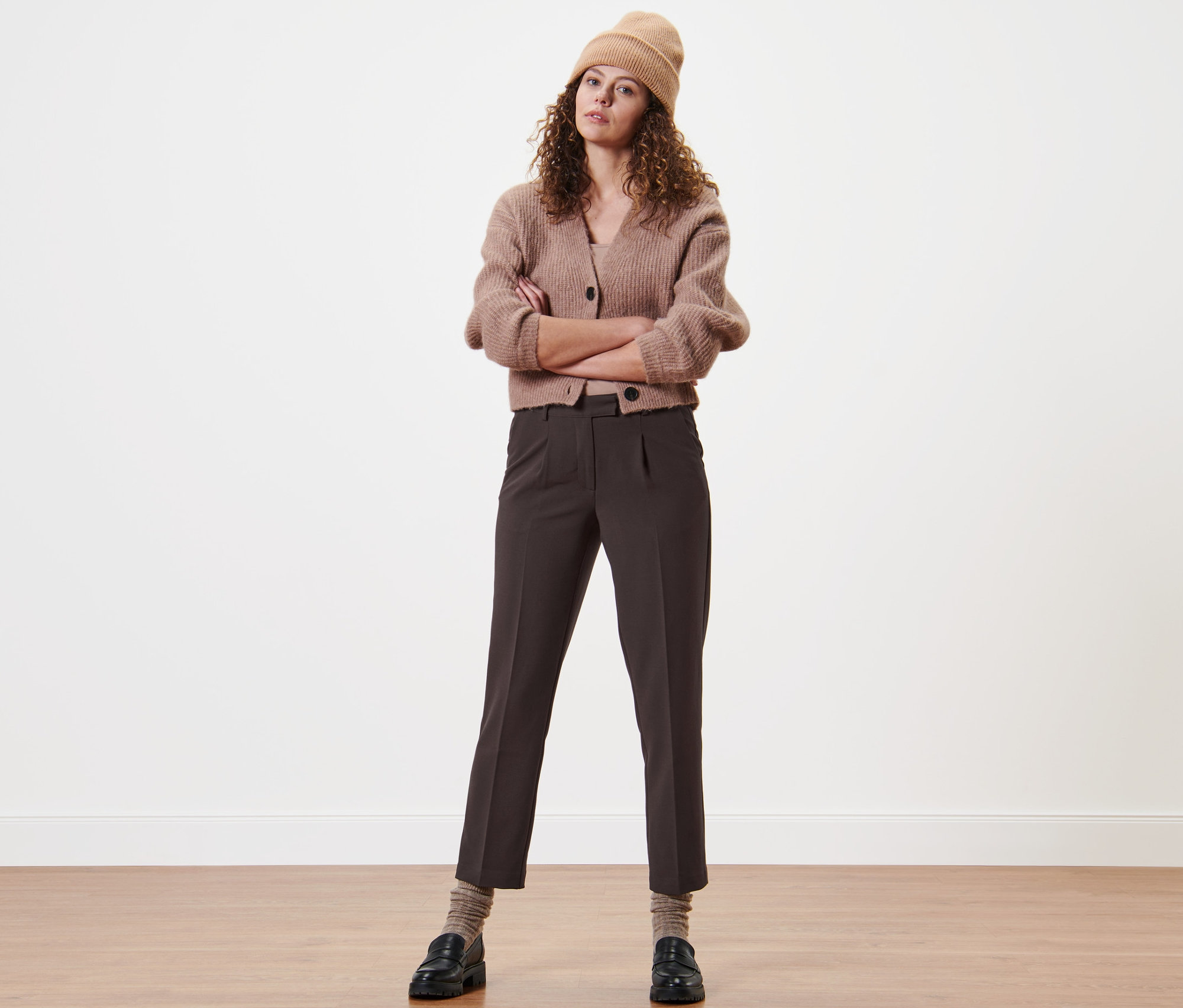 Une femme aux bras croisés porte un pantalon tissé à pinces, un cardigan et un bonnet.