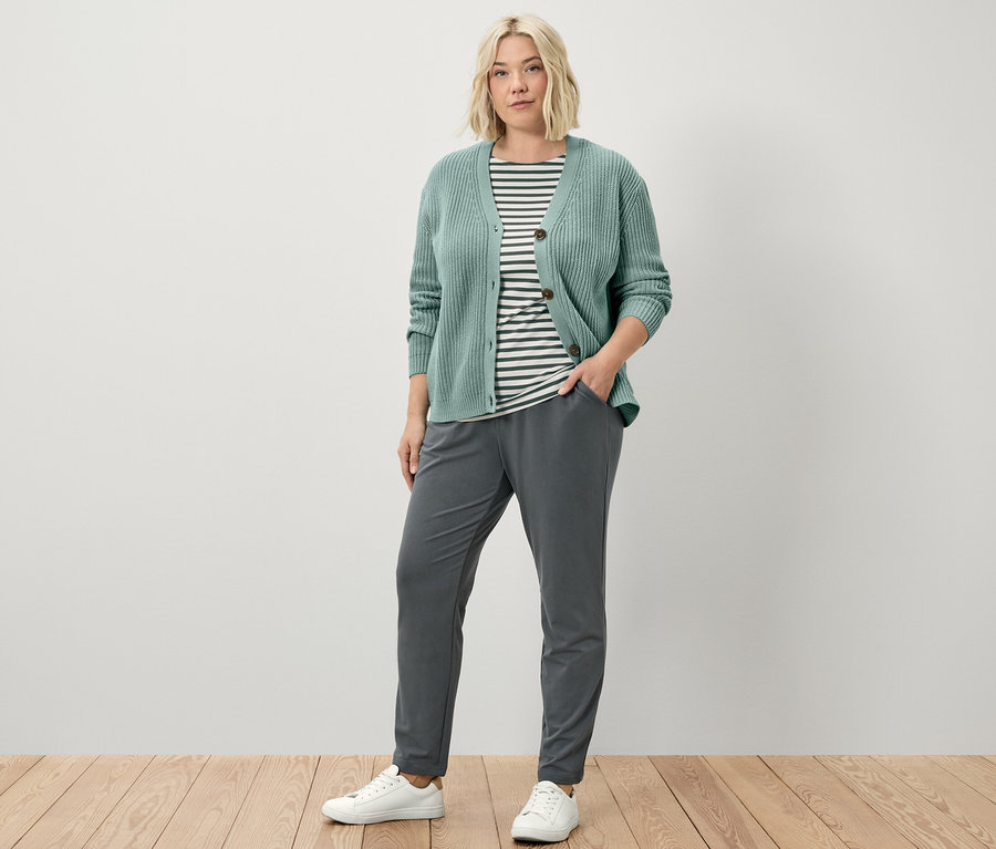 Une femme debout porte un pantalon de jogging, un cardigan en grosse maille sauge et un t-shirt à rayures vertes à manches courtes.