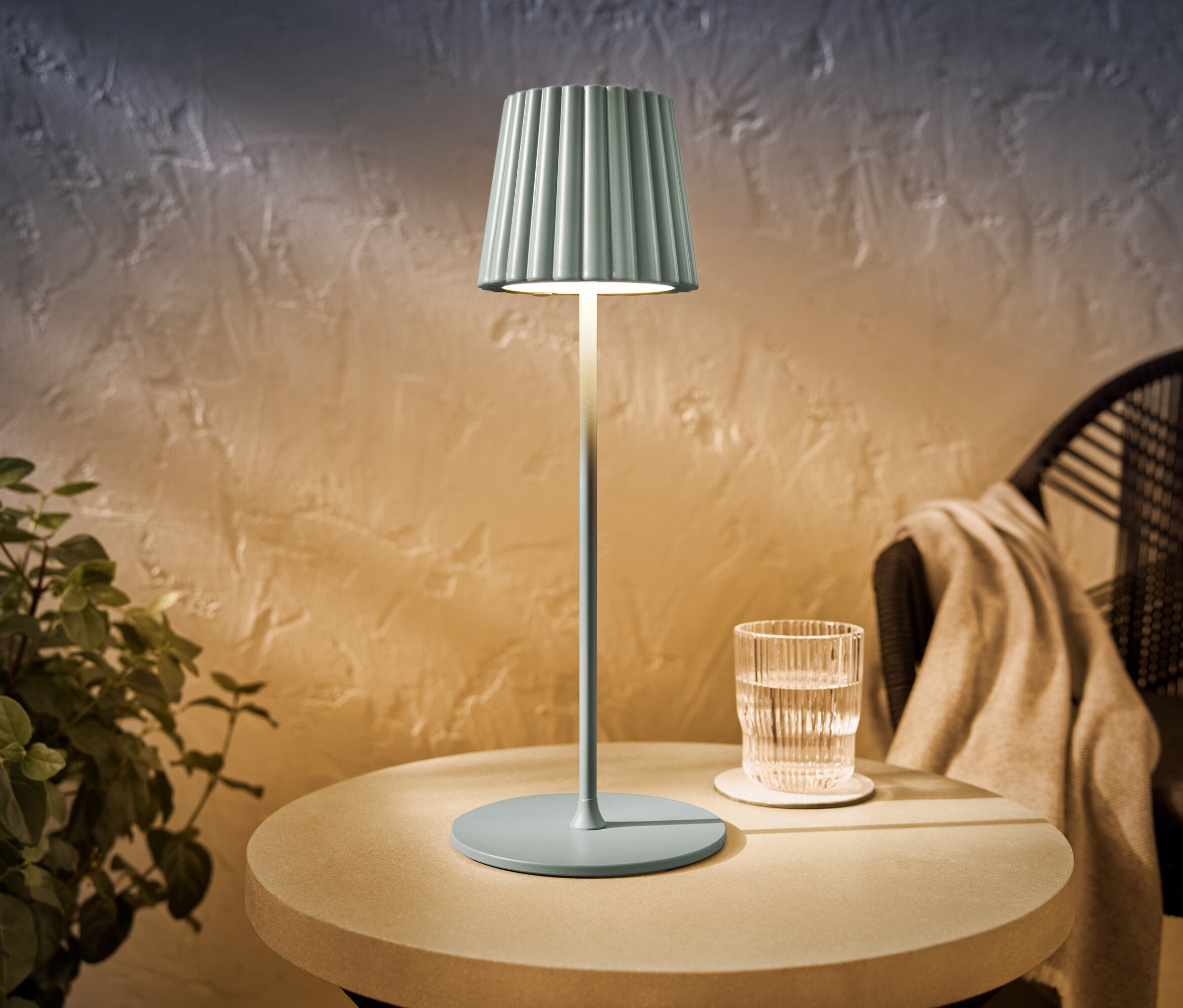 Une lampe de table d’extérieur à LED rechargeable sauge est posée sur une table ronde à côté d’un verre d’eau.
