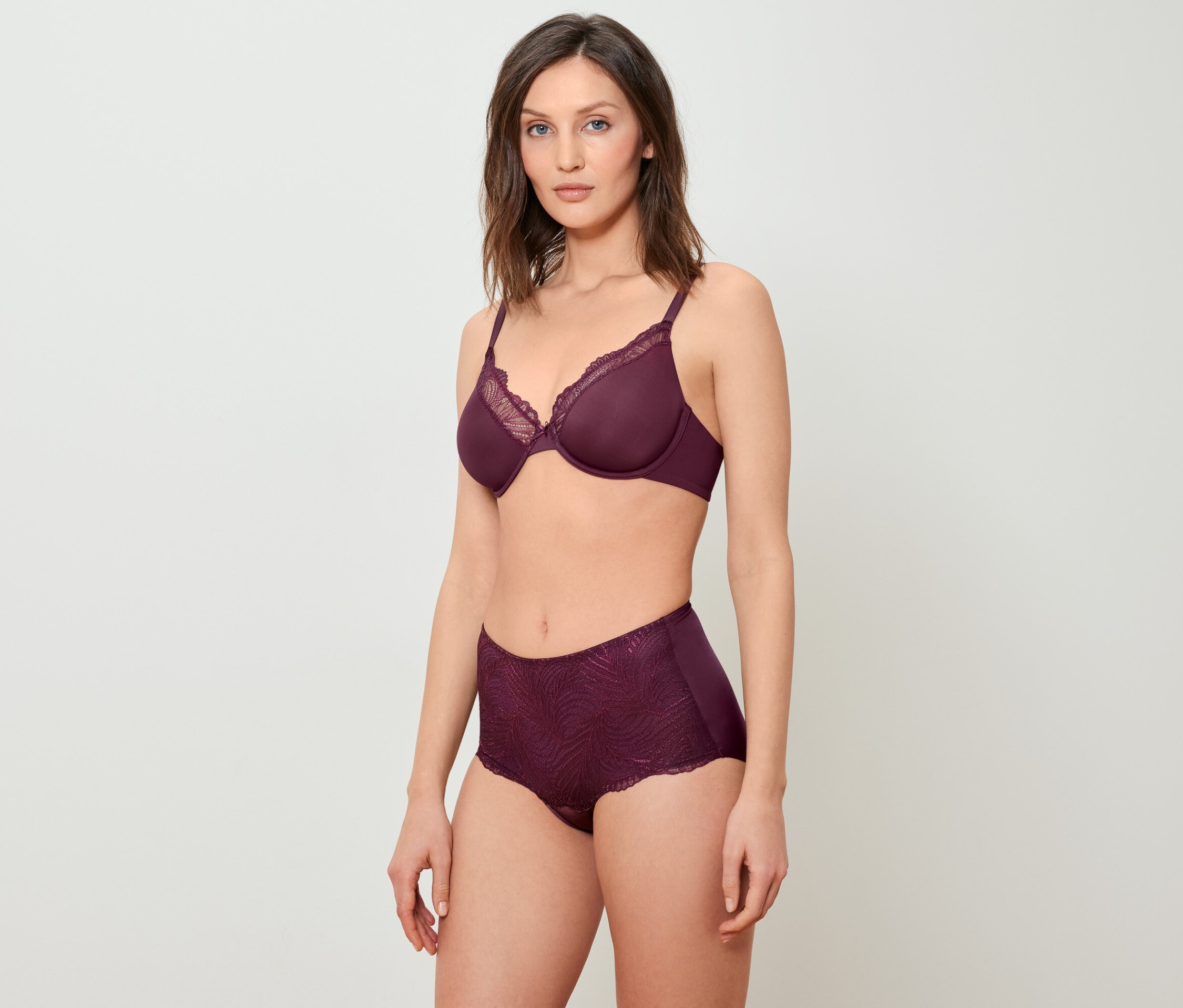 Une femme pose en soutien-gorge bordeaux avec de la dentelle et une culotte taille haute sur un fond blanc.