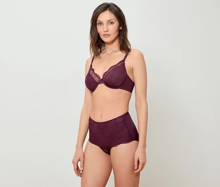 Une femme pose en soutien-gorge bordeaux avec de la dentelle et une culotte taille haute sur un fond blanc.
