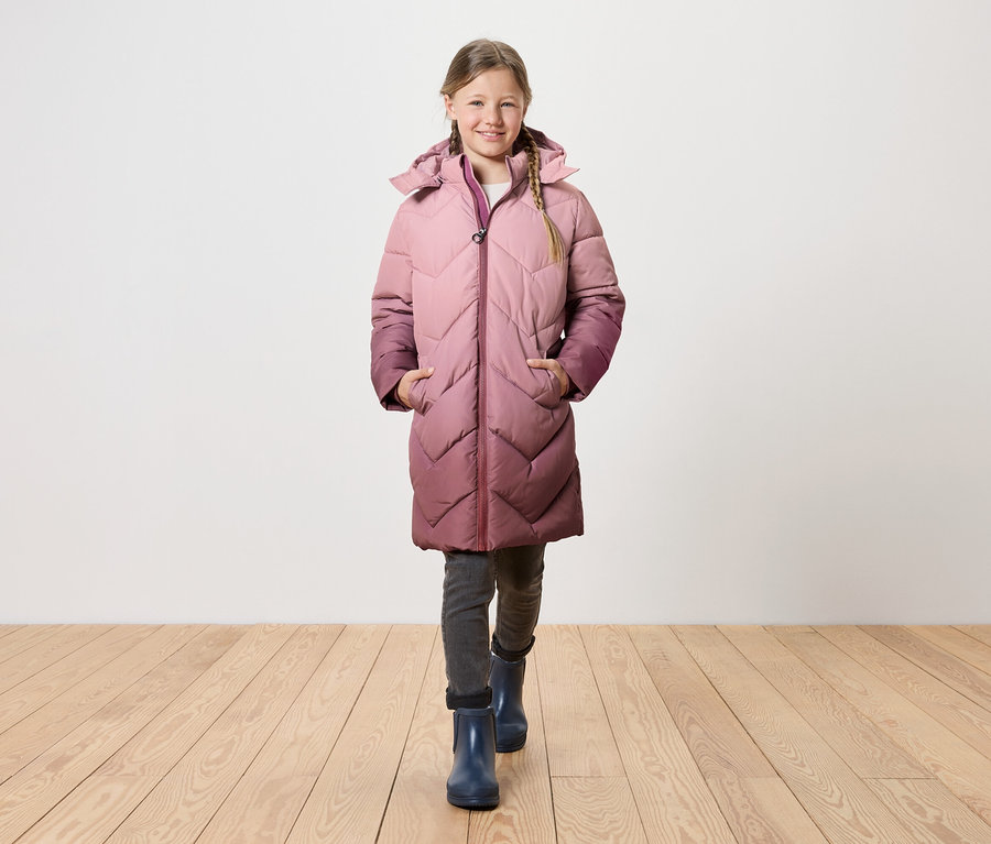 Une fille avec des tresses blondes marche sur un plancher en bois, portant un long manteau matelassé à capuche qui passe du rose au violet.