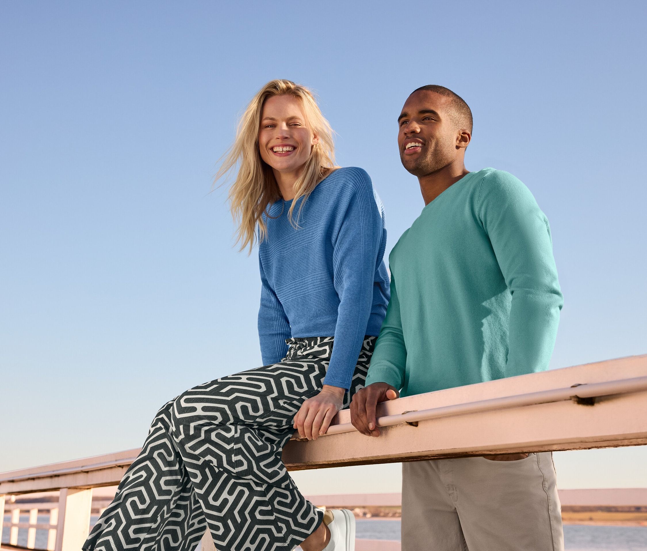 Une femme est assise sur une balustrade portant un pull en maille fine bleu et un pantalon imprimé à enfiler avec effet Crinkle. À côté d'elle se trouve un homme portant un pull en maille et un pantalon 5 poches.