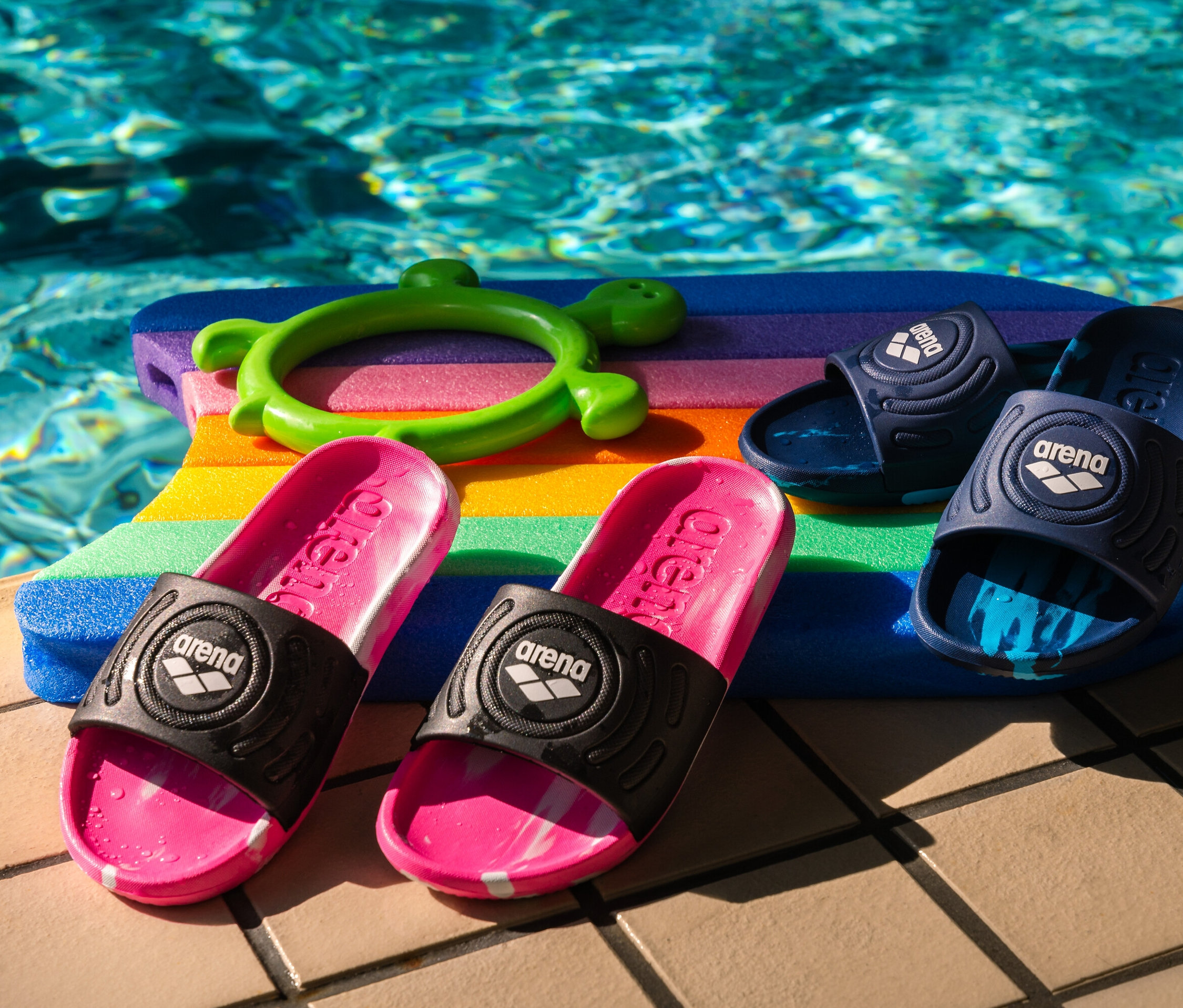 Deux paires de sandales de bain arena Junior Urban Active pour enfant, rose vif et bleu marine, posées sur une planche de natation colorée au bord de la piscine.