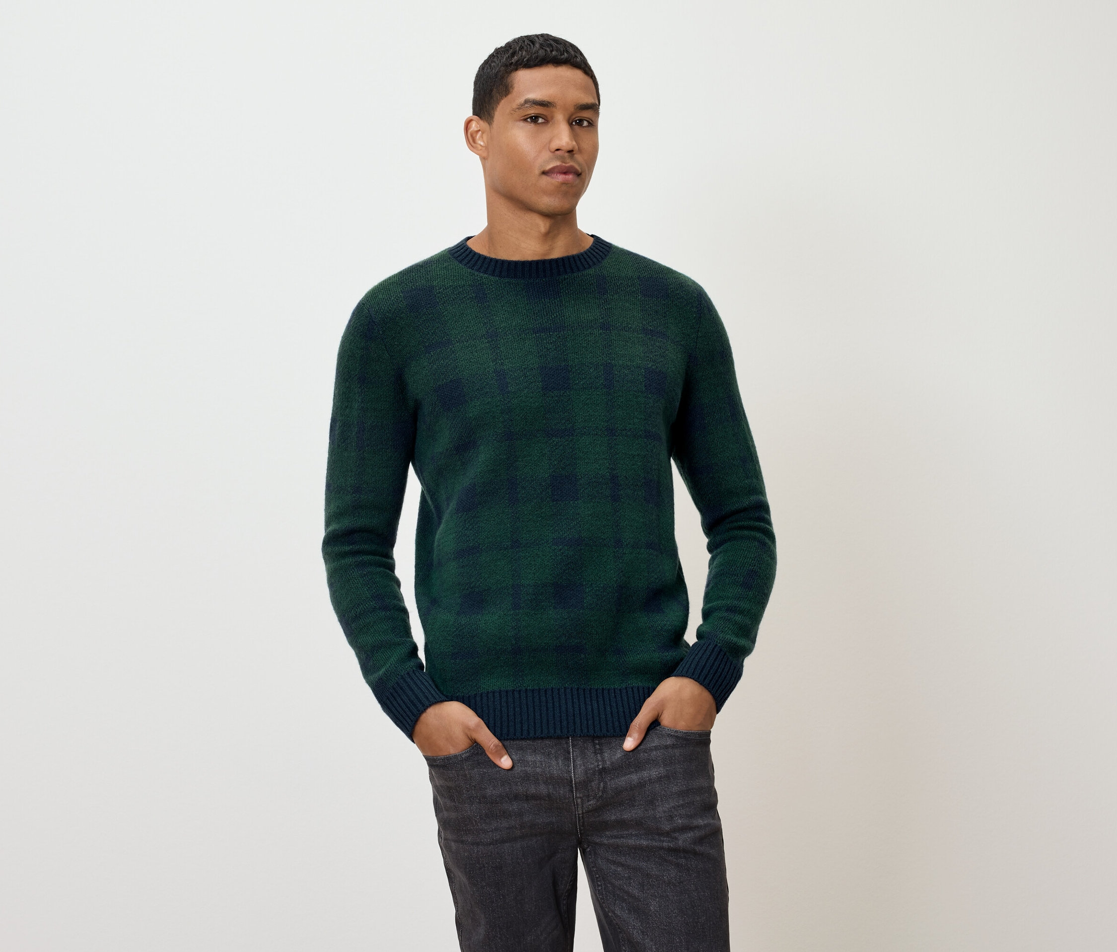 Un homme pose en pull à carreaux vert et jean gris.