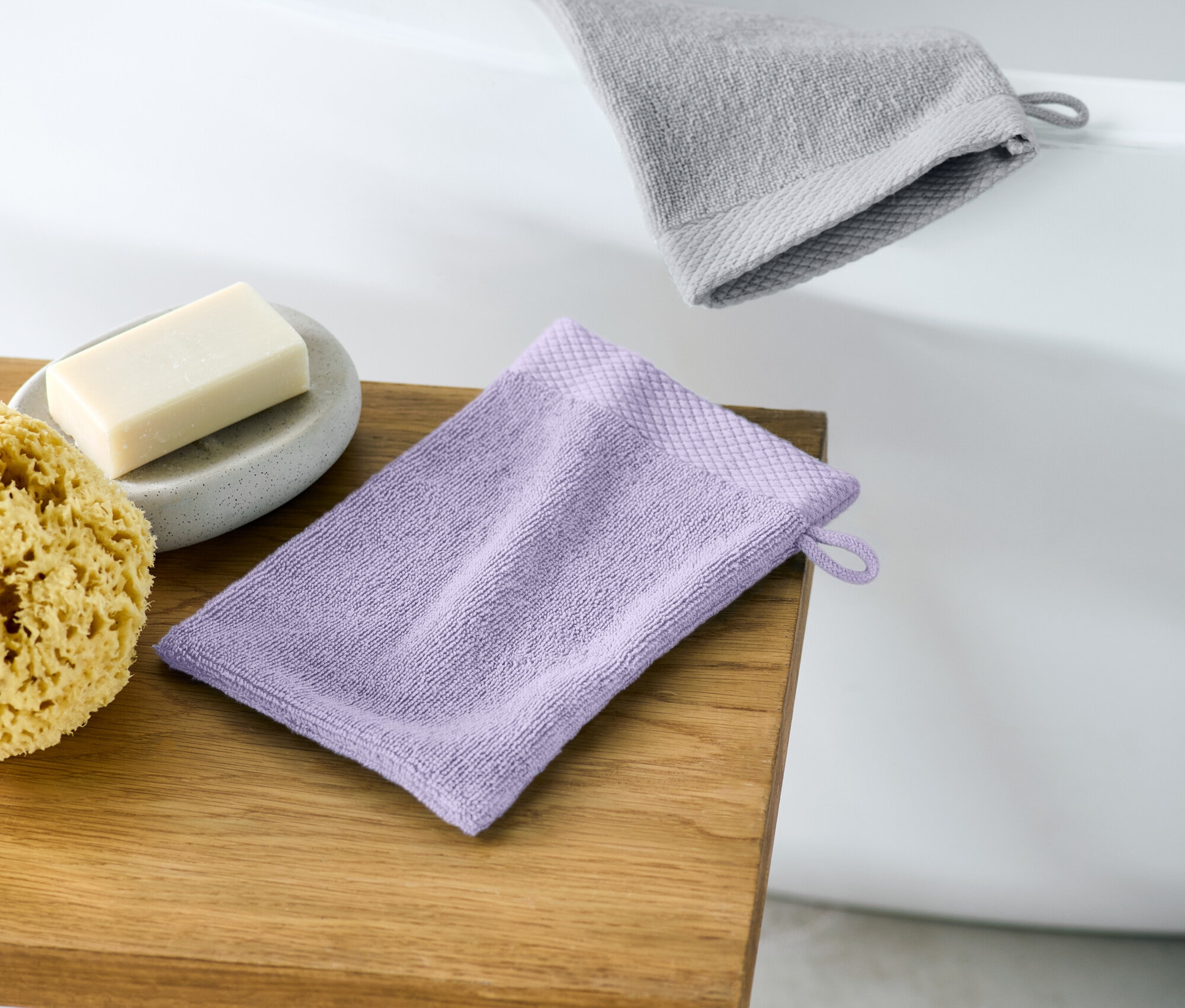Sur un tabouret en bois reposent un gant de toilette violet, à côté duquel se trouvent une éponge naturelle et un savon gris sur une soucoupe. À l'arrière-plan, on aperçoit le bord d'une baignoire et un gant de toilette gris.