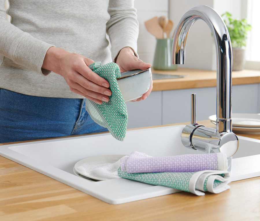 Une femme essuie un bol avec un torchon vert près d'un évier avec robinet et distributeur de savon.