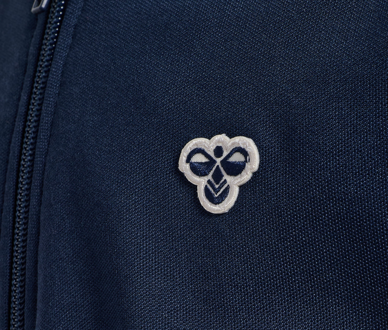 Gros plan sur une veste zippée bleue Hummel HMLJR Archive Reg Poly avec logo.