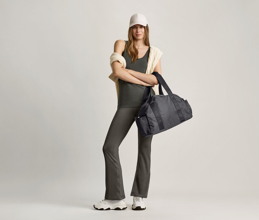 Une femme se tient debout, les bras croisés, et tient un sac de sport noir. Elle porte un haut vert olive et un pantalon gris.