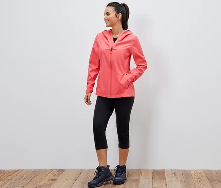 Une femme souriante pose en veste outdoor 2,5 couches corail, legging trois-quarts noir et chaussures de randonnée.