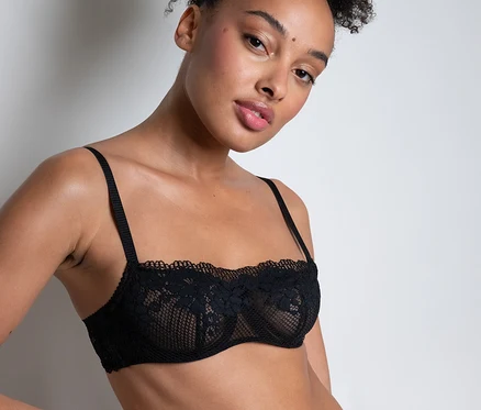 Une femme aux cheveux bouclés relevés porte un soutien-gorge bandeau « Nina » Passionata en dentelle noire et une culotte assortie, une main sur la hanche.