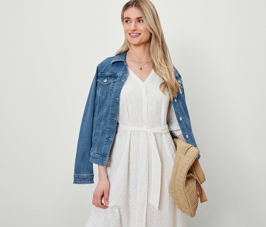Une femme porte une robe avec broderie ajourée et une veste en jean et porte un cabas aspect raphia.