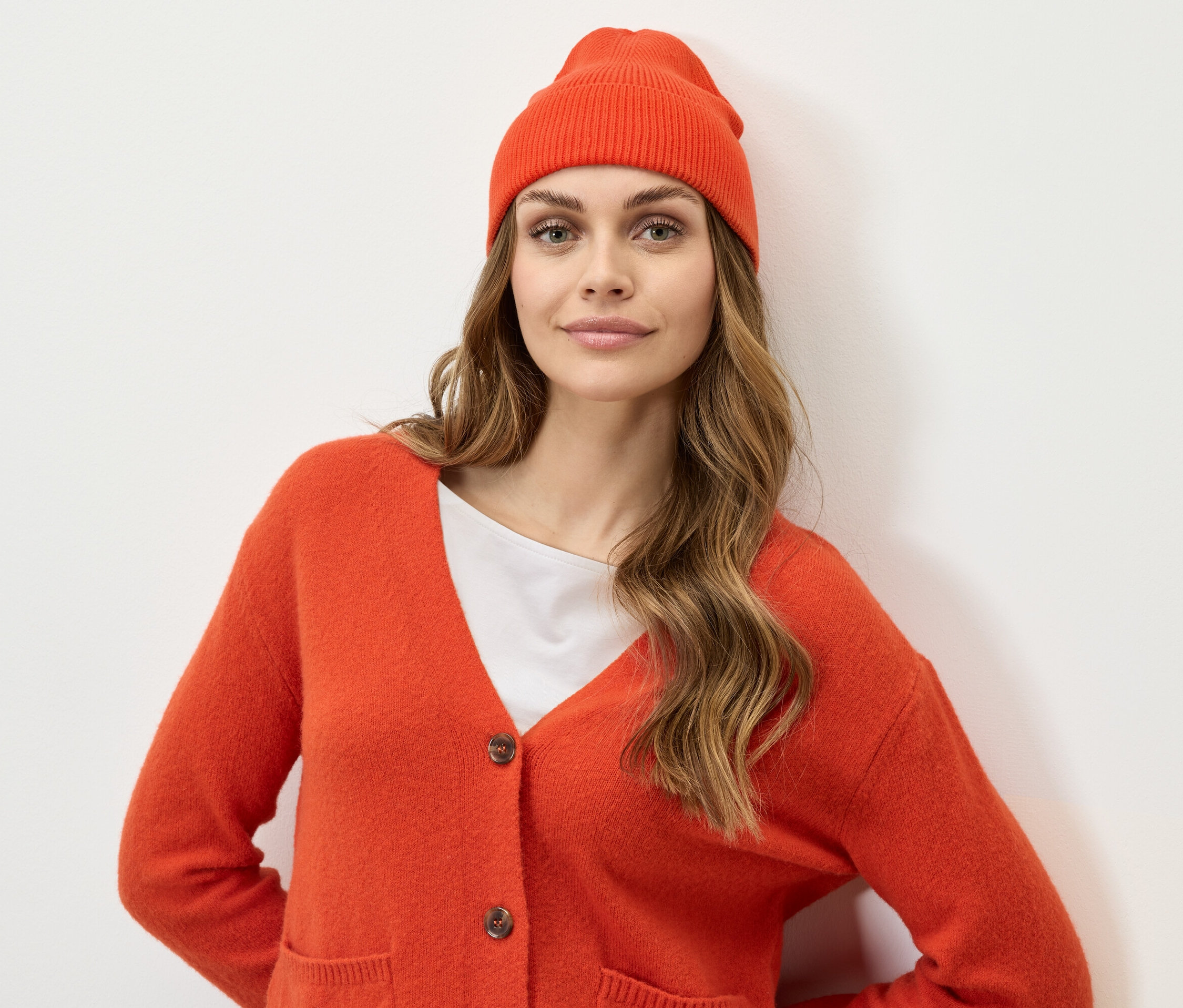 Une femme aux cheveux bruns, portant un bonnet et un cardigan orange, regarde la caméra.