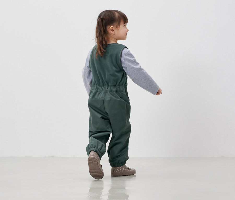 Fillette en salopette verte et chemise grise marchant sur un sol blanc.