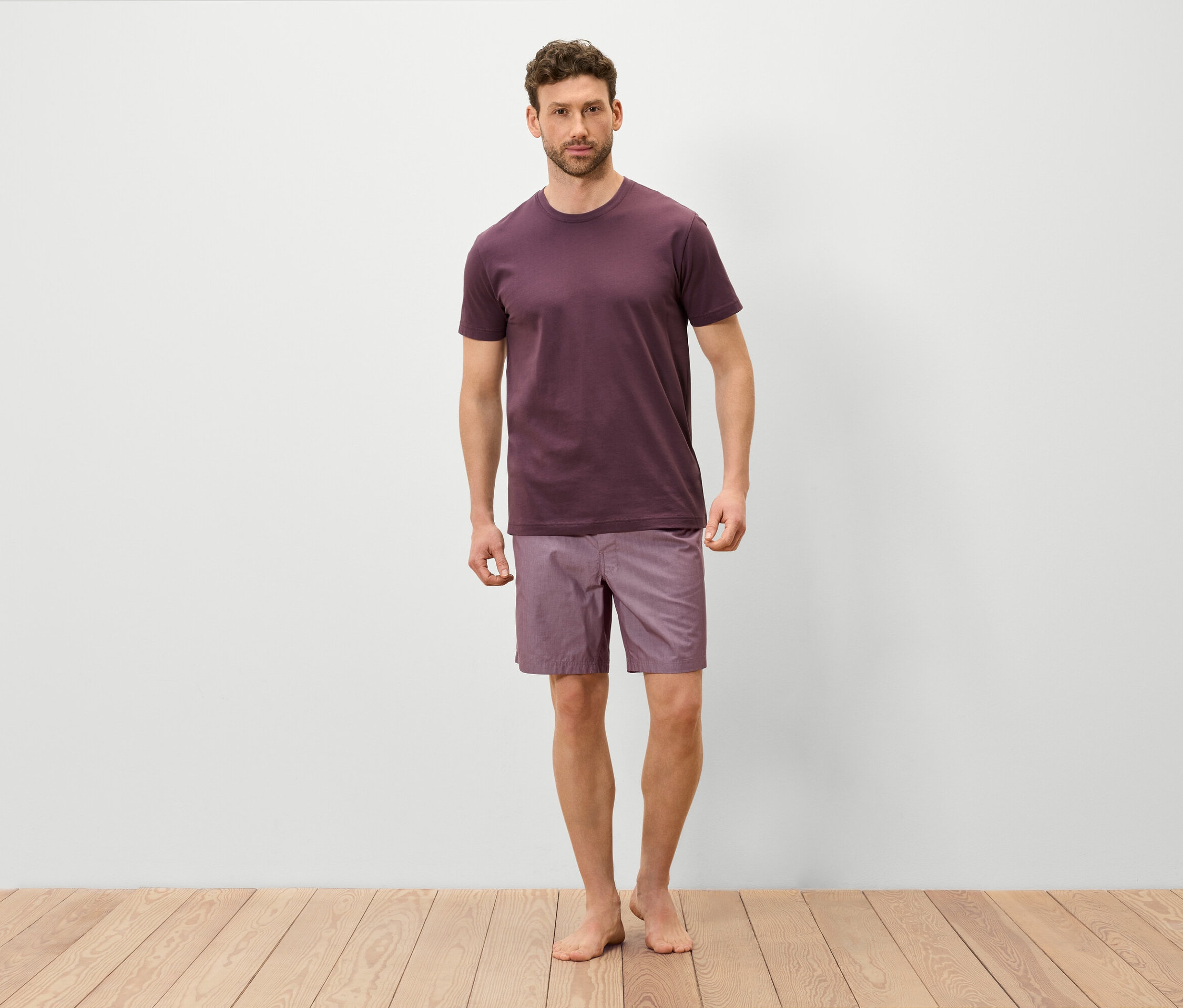 Un homme se tient pieds nus sur un plancher en bois, portant un t-shirt bordeaux et un short violet.