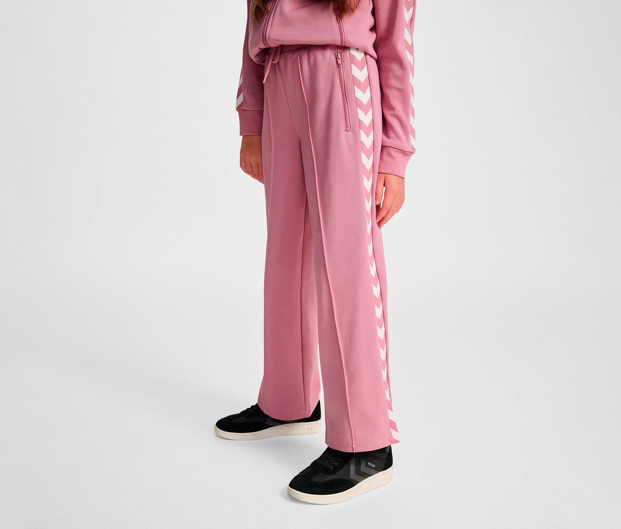 Une personne portant un pantalon large en polyester HUMMEL HMLJR Archive. Il est rose avec des bandes à chevrons blanches sur les côtés.