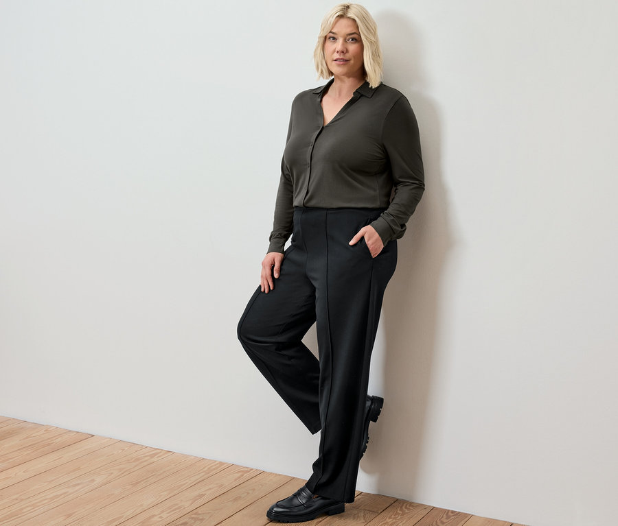 Une femme blonde appuyée contre un mur porte un pantalon en jersey noir avec passepoil et une blouse avec patte de boutonnage.