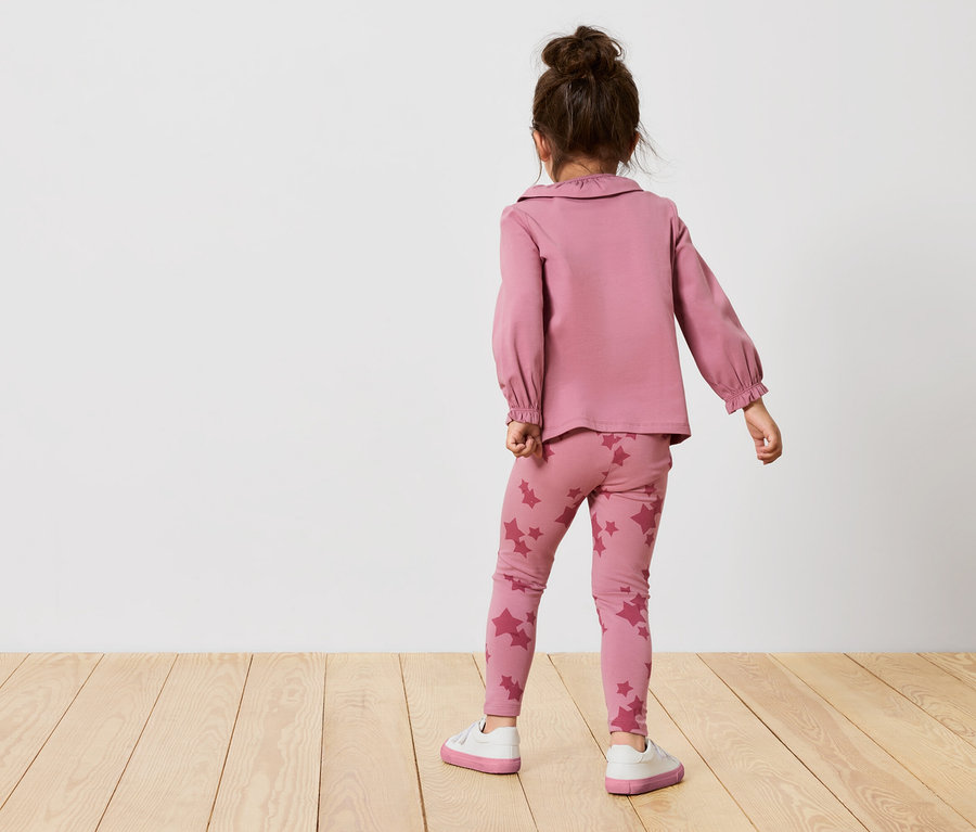 Une fille en ensemble rose à étoiles se tient dos tourné. Elle porte un haut rose à manches longues et un legging à étoiles.